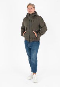 Giacca imbottita verde oliva con cappuccio, chiusura frontale con zip e due tasche laterali; abbinata a jeans blu e sneakers bianche.