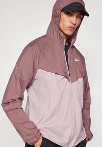 Kerge kapuutsiga jakk kahes toonis; ülemine osa roosakas ja alumine hele roosa, esiosas tõmblukk ja logo.