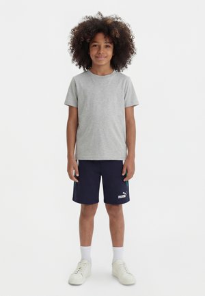 Kind mit lockigem Haar, trägt ein graues T-Shirt, marineblaue Puma-Shorts, weiße Socken und weiße Turnschuhe, steht vor einem schlichten Hintergrund.