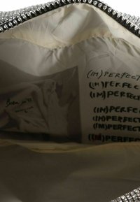 Bolso negro forrado con tela crema, con herrajes plateados con cuentas y texto impreso que dice "(IM)PERFECT" en negro en el interior.