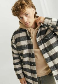 Camicia di flanella a quadri nera e crema, con grandi tasche e bottoni a pressione, indossata sopra un hoodie beige. Jeans di denim visibili sotto.