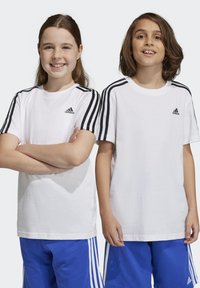 Dos niños que llevan camisetas blancas de Adidas con acentos en negro en las mangas, de pie en pantalones cortos deportivos azules.