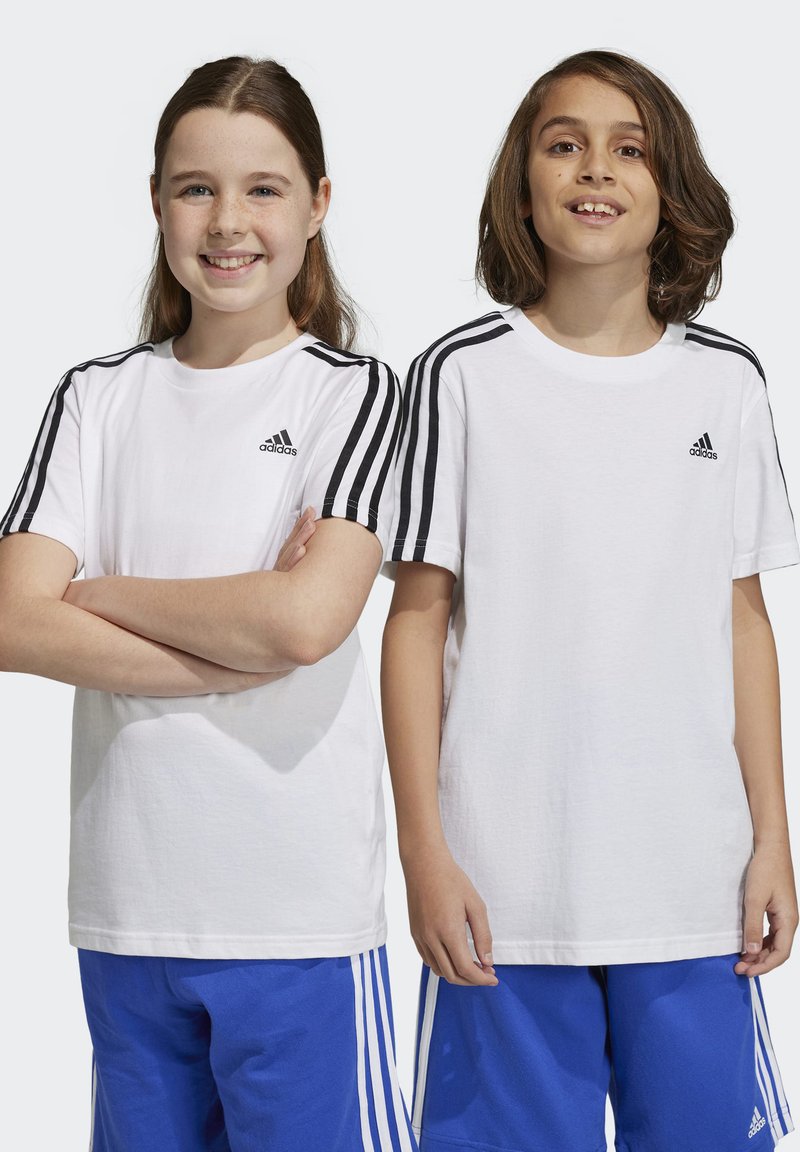 Dos niños que llevan camisetas blancas de Adidas con acentos en negro en las mangas, de pie en pantalones cortos deportivos azules.