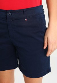 Námořnické modré bavlněné kraťasy s knoflíkovým zapínáním, poutky na opasek a bočními kapsami. Vyznačují se malým červeným a bílým akcentem poblíž pasu.