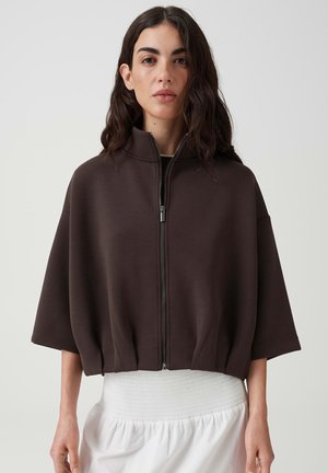 Frau mit dunklem, welligem Haar, die ein lockeres dunkelbraunes Zip-up-Oberteil mit breiten kurzen Ärmeln und hohem Kragen über einem weißen Rock trägt.