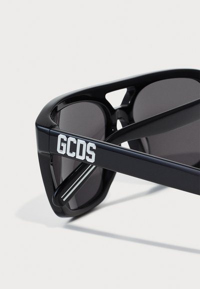 Gafas de sol negras con acabado brillante, que presentan un diseño angular fuerte, lentes oscuros y una fuerte marca blanca en la patilla.
