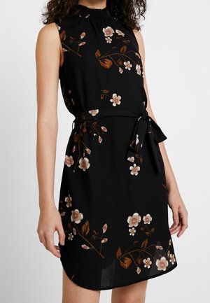 Robe noire sans manches avec des motifs floraux marron et beige, col montant et ceinture à la taille, portée par une personne debout.