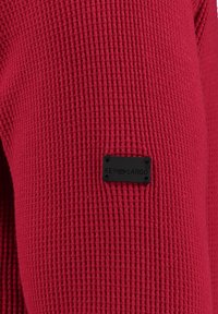 Vêtement en tissu texturé rouge avec un design côtelé et une étiquette noire portant le texte "KEY LARGO" attachée à la manche.