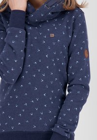 Sudadera azul marino hecha de una tela suave con cuello tipo cowl, que presenta un patrón moteado en azul claro. Incluye un parche de cuero con el logo en la manga.