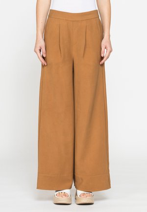 Donna che indossa pantaloni marroni a vita alta e gamba larga con pieghe frontali e sandali bianchi con zeppa, in piedi davanti a uno sfondo chiaro e uniforme.