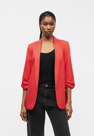 Femeie purtând un sacou roșu aprins peste un top negru și blugi întunecați cu talie înaltă, stând în fața unui fundal simplu, luminos.