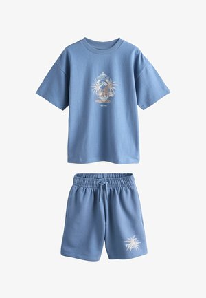 Blåt sæt med kortærmet skjorte og shorts med tropiske palmetræsgrafikker og teksten "Tropical" på forsiden af skjorten og venstre ben af shortsene.