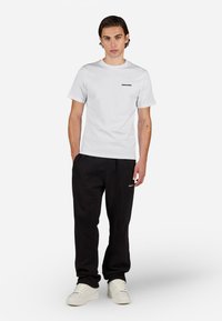 Biela tričko s krátkym rukávom a čiernym logom v kombinácii s čiernymi teplákmi. Model má na sebe biele tenisky a stojí na jednoduchom pozadí.