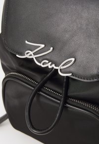 Black backpack made of synthetic material, featuring a silver metal logo, zippered pocket, and a rounded top handle.