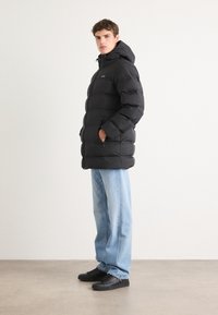 Zwarte pufferjack met capuchon, voorzien van een gewatteerd ontwerp, ritszakken en zwarte sneakers. Gecombineerd met lichtblauwe straight-leg jeans.