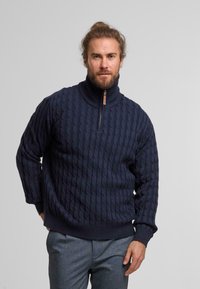 Maglione in lana navy a trama cable knit con colletto alto, chiusura a metà zip e orlo a coste. Il tessuto testurizzato crea un motivo di onde intrecciate.