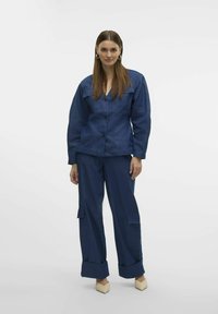Completo giacca e pantaloni in denim blu scuro. La giacca presenta una chiusura con bottoni e due tasche sul petto. I pantaloni hanno gambe larghe e tasche cargo.
