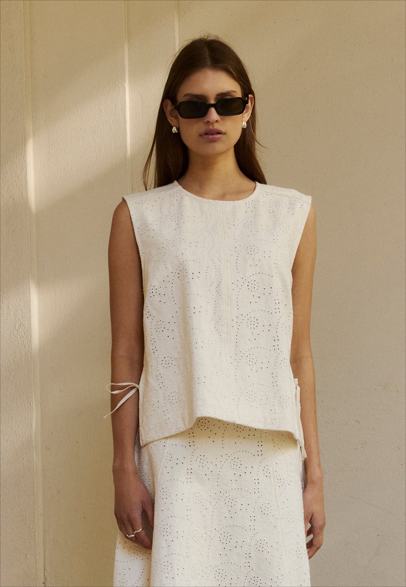 Femme portant un haut sans manches blanc en œillets et une jupe assortie, avec des lunettes de soleil noires rectangulaires et des boucles d'oreilles en perles, debout contre un mur beige.