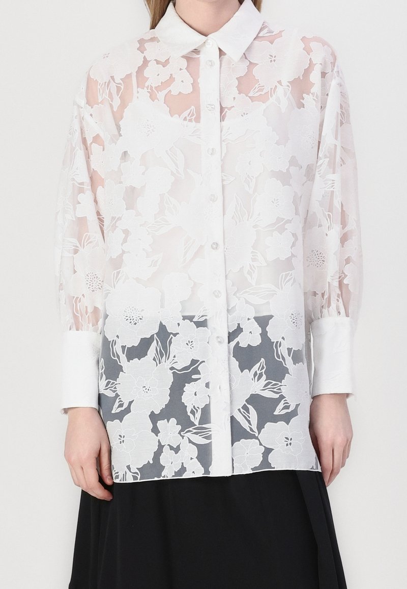 Femme portant une blouse blanche transparente à boutons avec des motifs en dentelle florale sur une jupe noire, devant un fond uni.