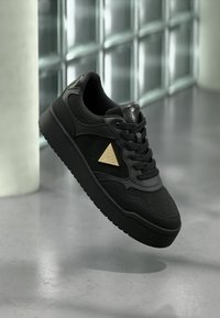 Mustad musta värvi sneaker, millel on reljeefne muster ja kuldne metallist logo. Omab ümmargusi paelu, tekstureeritud pinda ja paksu, tahket tallat.