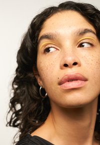 Nahaufnahme einer Person mit lockigem Haar und Sommersprossen, die gelinertes Augen-Make-up, kreisförmige Ohrringe und eine natürliche Lippenfarbe trägt, vor einem neutralen Hintergrund.