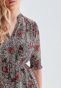 Vestido cruzado de tela ligera con una base beige y estampado de leopardo negro, además de patrones de paisley en rojo y verde. Mangas con elástico.