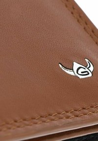 Surface en cuir marron avec une couture soignée et un petit emblème argenté en forme de casque stylisé à cornes avec un « G » à l'intérieur.