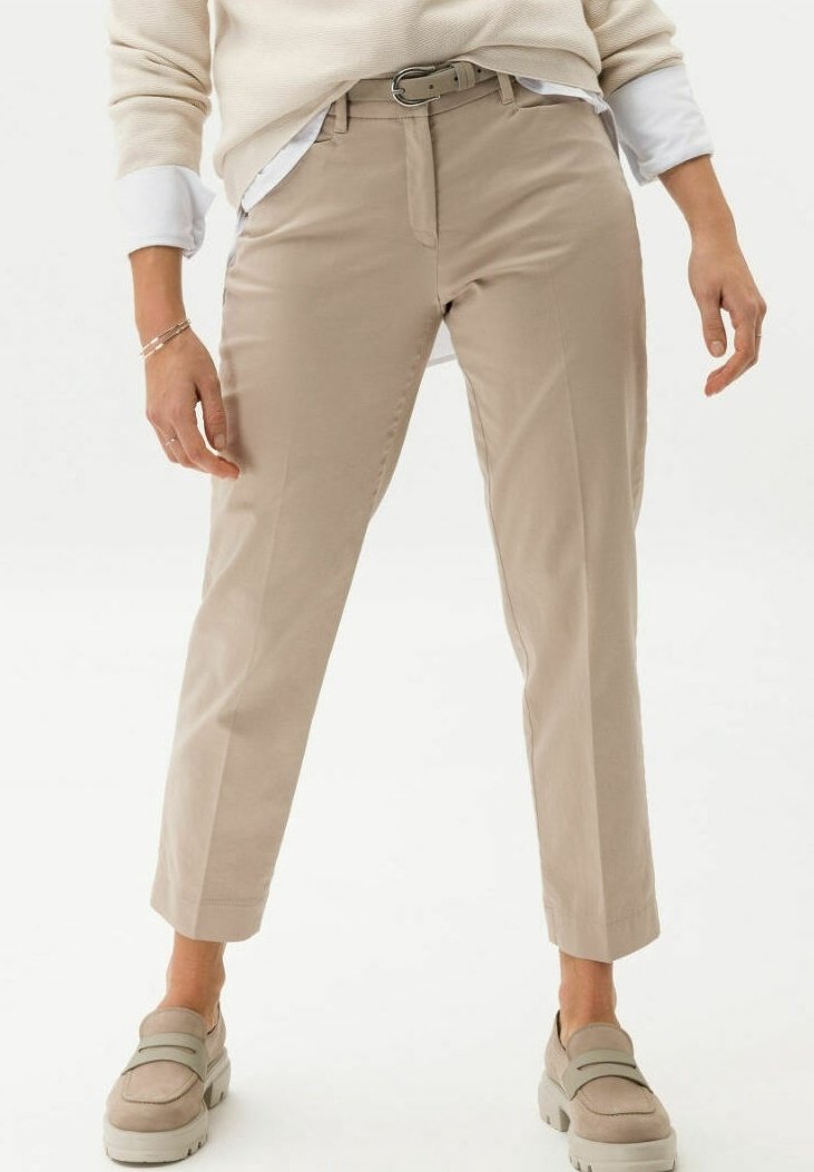Personne portant un pantalon court beige clair avec une ceinture, des mocassins beiges, une chemise blanche et un pull crème avec les manches retroussées.