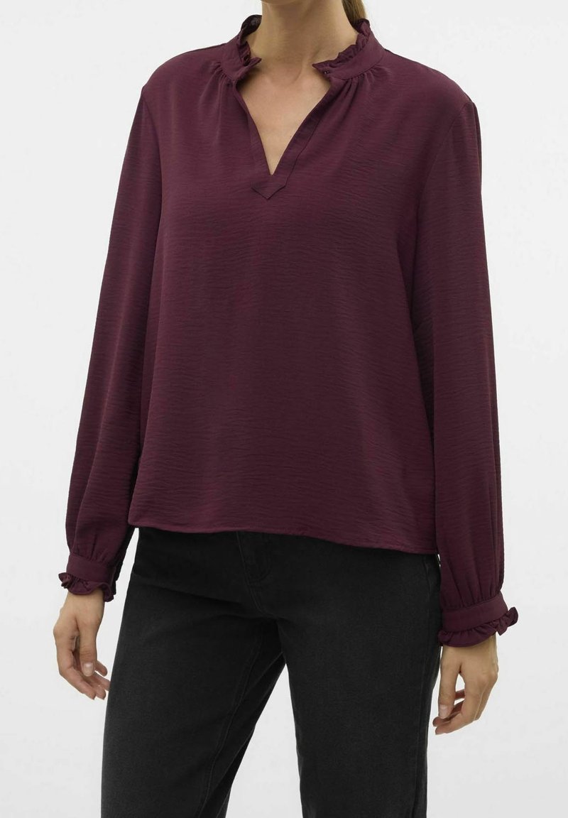 Femme portant un chemisier violet à manches longues avec col et poignets volantés, associé à un pantalon noir sur un fond uni.