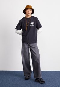 Mężczyzna w brązowym kapeluszu typu bucket, czarnej koszulce z nadrukiem na białej długiej koszuli, szerokich szarych dżinsach i czarnych sneakersach stoi na niebieskim dywanie.