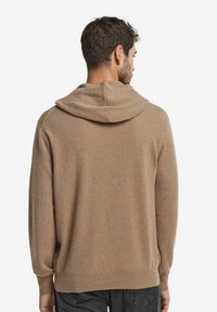 Mann, der von hinten zu sehen ist, trägt einen hellbraunen Kapuzenpullover und dunkle Jeans vor einem schlichten weißen Hintergrund.