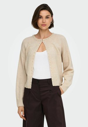 Femme portant un cardigan beige en maille avec des ornements en perles sur un haut blanc et un pantalon taille haute marron foncé, debout avec une main dans la poche.