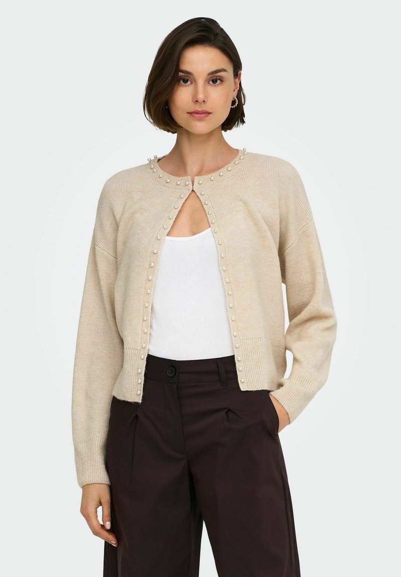 Femme portant un cardigan beige en maille avec des ornements en perles sur un haut blanc et un pantalon taille haute marron foncé, debout avec une main dans la poche.