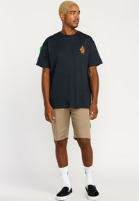 T-shirt en coton noir avec un petit logo orange sur la poitrine, associé à un short beige. Le modèle porte des chaussures noires à enfiler et des chaussettes blanches.
