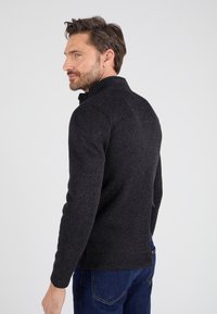 Suéter negro texturizado con cuello alto, diseño ajustado y mangas largas, combinado con pantalones vaqueros azul oscuro. Estilo simple y funcional.