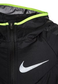 Kurtka Nike wykonana z lekkiego czarnego materiału o teksturowanym wykończeniu, z fluorescencyjno-zielonymi akcentami w okolicy kołnierza i zamka.