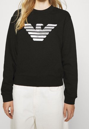 Sudadera negra con un diseño recortado, que presenta un logo de águila blanca. La tela parece suave con puños y dobladillo acanalados.