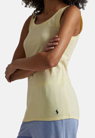 Femme aux bras croisés portant un haut sans manches jaune pâle et un pantalon bleu clair, sur fond uni.