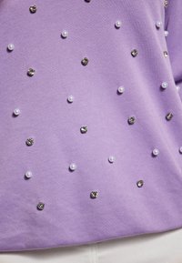 Felpa lilla realizzata in materiale morbido, presenta perle bianche e gemme metalliche sparse come dettagli, superficie testurizzata con finitura liscia.