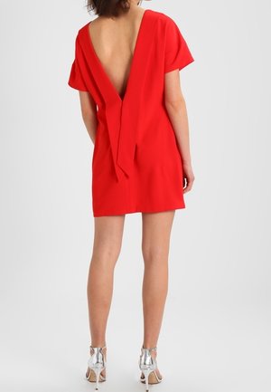 Rotes Kleid mit tiefem V-Ausschnitt hinten, kurzen Ärmeln und einem gebundenen Detail am Rücken. Glatte Textur, knielang, kombiniert mit silbernen High Heels.