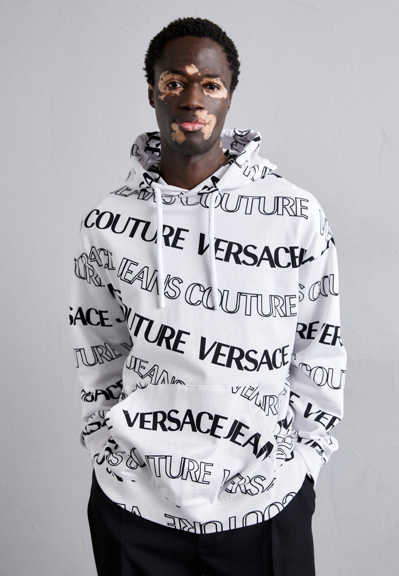 Hoodie blanc avec un imprimé noir répétitif de "Versace" et "Jeans Couture". Comprend des cordons de serrage et une poche frontale. Matériau doux.