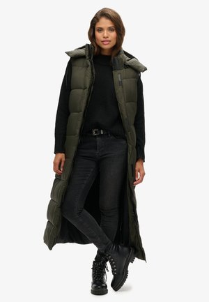 Un long gilet rembourré vert avec une capuche, présentant une fermeture éclair et des fentes sur les côtés, associé à un pull noir et un jean foncé. Des bottes noires à lacets.
