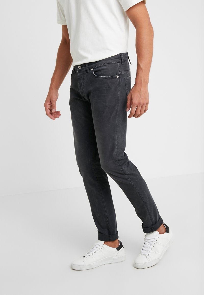 Homme portant un jean noir slim-fit retroussé aux chevilles, des baskets blanches et une chemise blanche à manches courtes, debout sur un fond blanc.