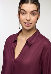 Camicia satina bordeaux con vestibilità rilassata, colletto button-down e profonda scollatura a V. Presenta una texture liscia e una lucentezza sottile.