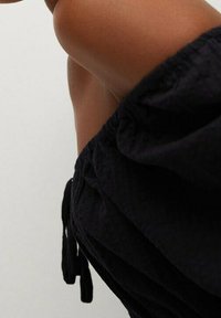 Schwarzes textiles Kleidungsstück mit einem elastischen Bund und Kordeln, das eine Nahaufnahme von Stofffalten auf der Haut zeigt.
