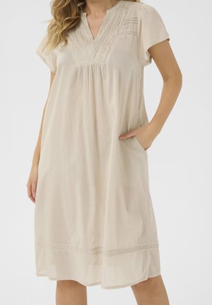 Mujer vistiendo vestido beige hasta la rodilla con mangas cortas, escote en V plisado, ribete de encaje y bolsillos laterales, de pie con una mano en el bolsillo.