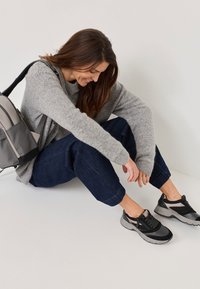 Schwarze und graue Sportschuhe mit Mesh- und Lederelementen. Kombiniert mit dunklen, locker sitzenden Jeans und einem grauen Pullover, sitzt auf einer hellen Oberfläche.
