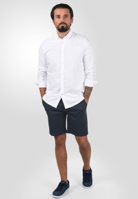 Chemise blanche à boutons, manches retroussées, associée à un short bleu marine. Portée avec des baskets bleu marine. Textures lisses, coupe décontractée et design simple.
