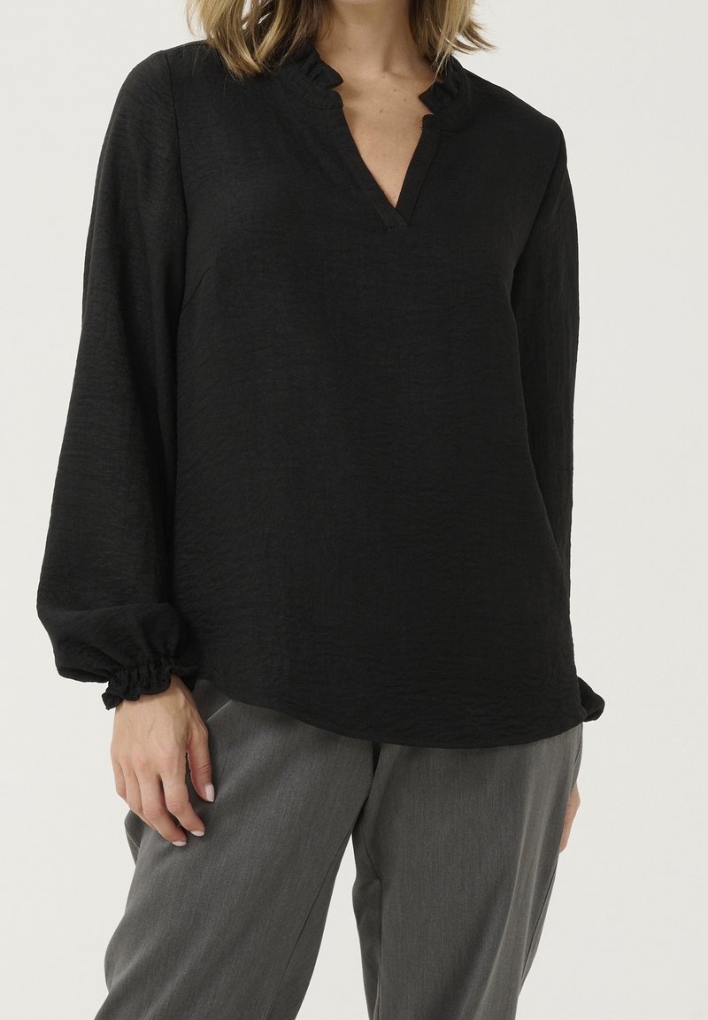 Top negro de manga larga con un acabado texturizado, que presenta un escote en V y un cuello con volantes. Combinado con pantalones grises, destaca por su estilo relajado.
