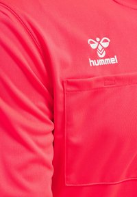 Lyserød sports skjorte lavet af glat stof, med en frontlomme og et hvidt Hummel-logo, samt synlige syningsdetaljer.